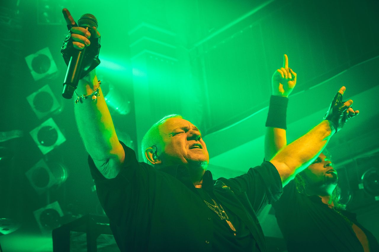 Udo Dirkschneider reißt in grünem Licht beide Arme nach oben, Mikrofon in der Hand