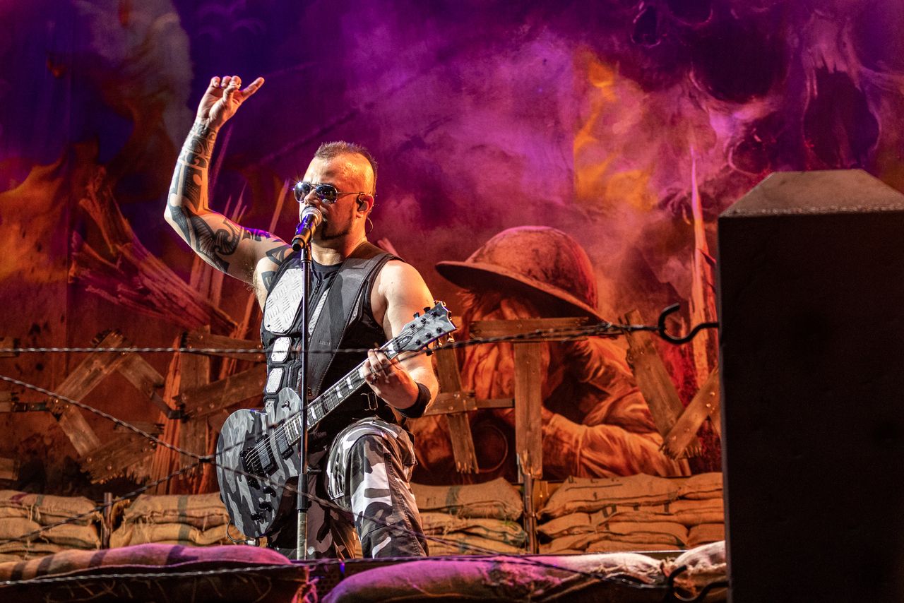 Sabaton-Sänger kniet mit Gitarre vor der Schützengraben-Kulisse in Orange und Violett