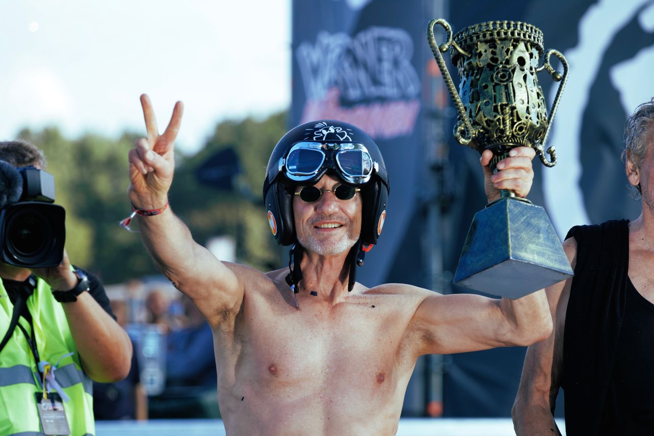 Sieger mit Motorradhelm und Fliegerbrille reckt jubelnd den Pokal in die Höhe und zeigt das Victory-Zeichen